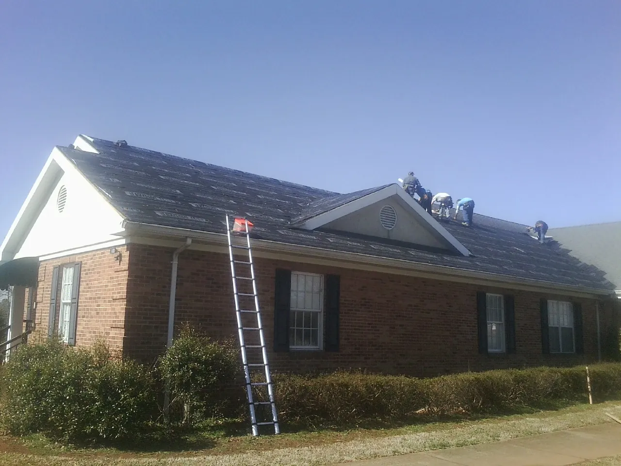 Crew tearing shingles off a brick ranch roof with a ladder set — Emergency Roof Repair work by our crew in Mineral Springs